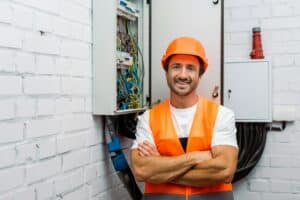 Handsome electrician with crossed arms smiling at camera near electric panel